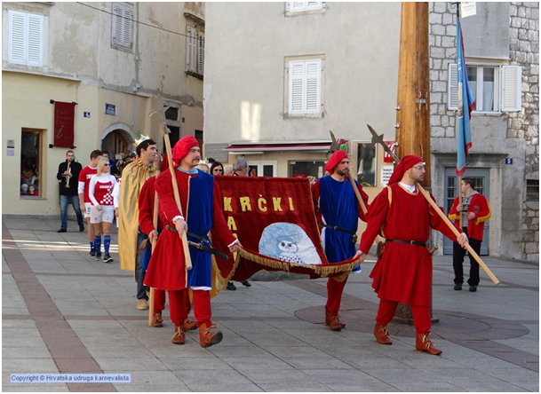 Men dressed in traditional costumes carry spears and a flag at the carnival.