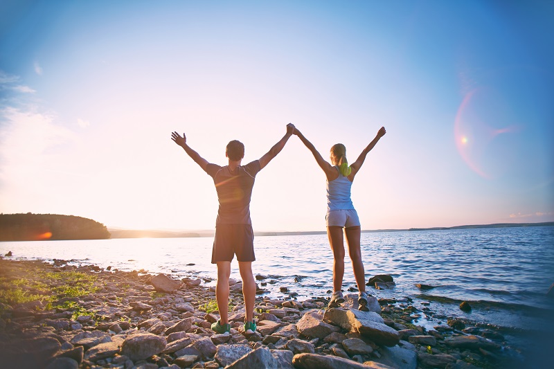 Muškarac i žena s podignutim rukama stoje na obali okrenuti prema moru.