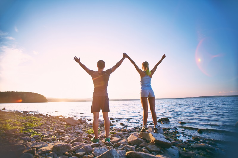 A man and a woman with raised hands stand on the shore facing the sea.